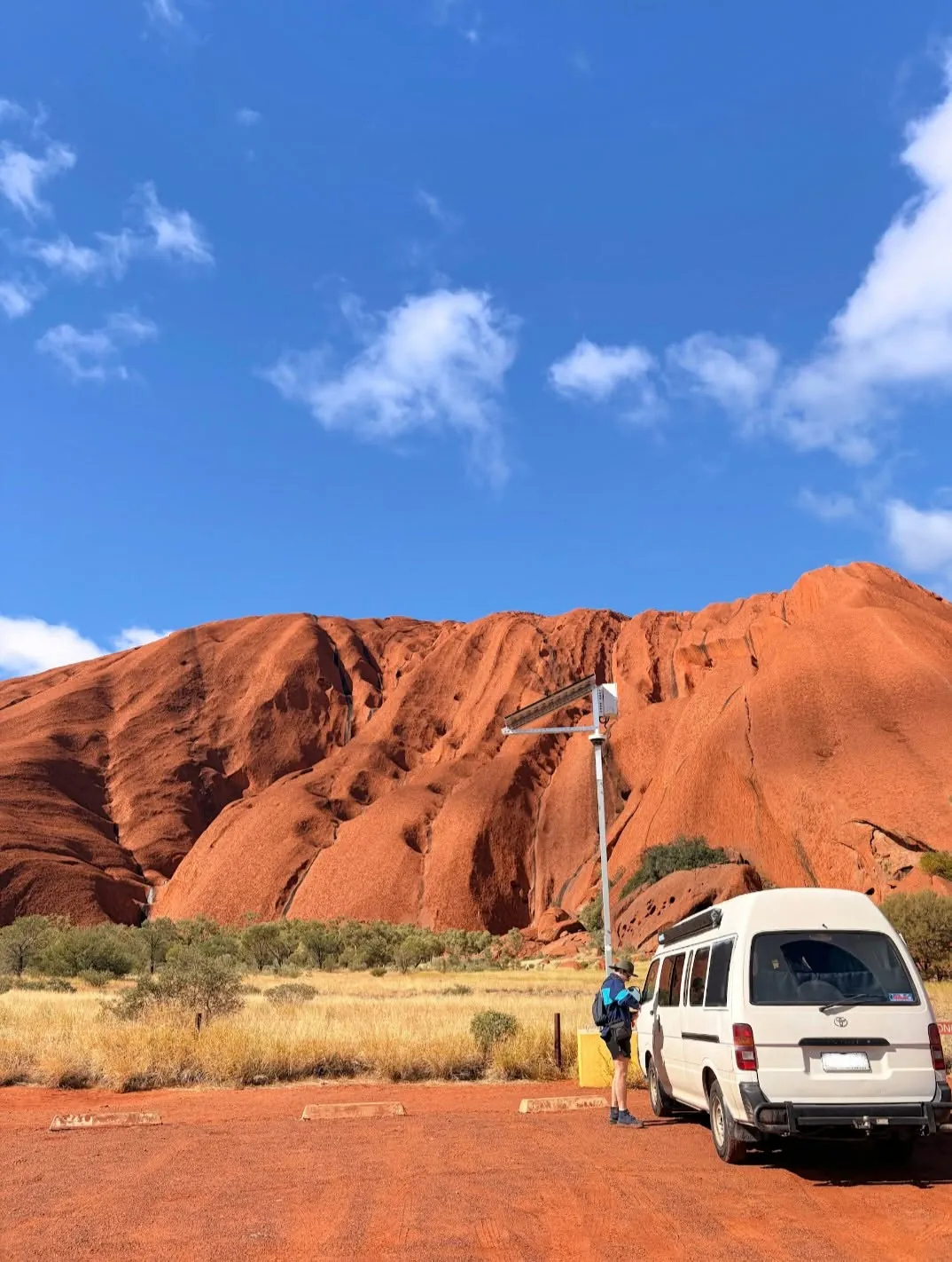Hank parked in front of Uluru's red rock face under a blue sky